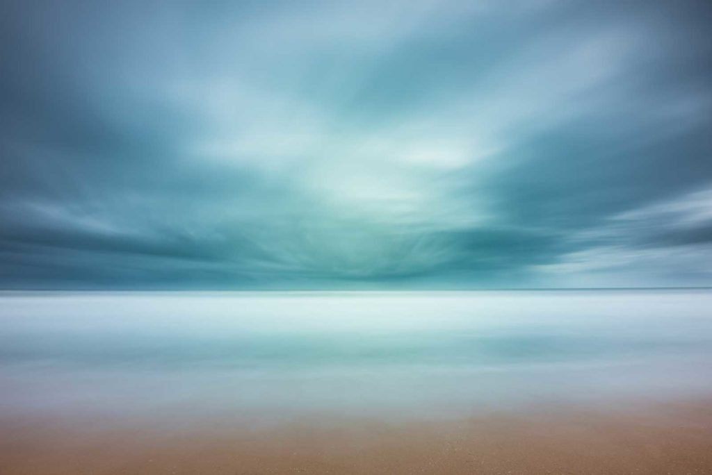 wide view of the ocean with beach in the foreground, blue strormy cloud cover creating an abstract view by the long exposure effect